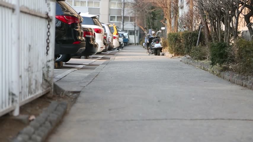 A low-angle view of a residential alleyway with parked cars along the sidewalk and a scooter parked in the distance