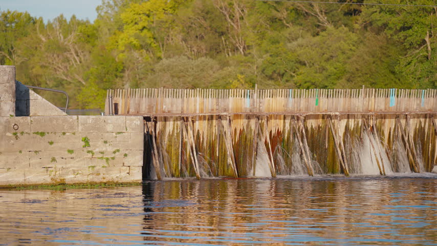 Water flows through a wooden needle dam structure beside a stone wall in a river setting