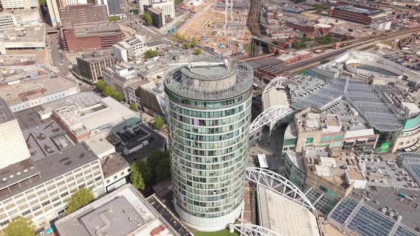Smooth aerial orbit around a modern cylindrical high-rise building in Birmingham city centre, revealing surrounding railway infrastructure, urban architecture and the dense city landscape.