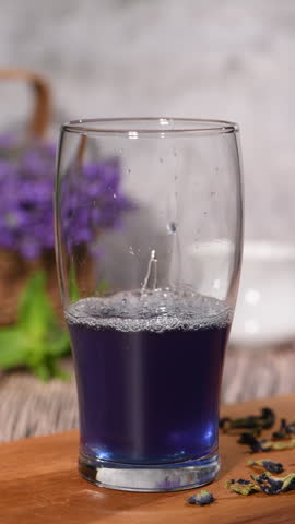 Clear ice cubes are added to a tall glass filled with blue butterfly pea tea, gradually cooling this vibrant floral drink. Close-up of ice falling into the drink. Part 3 of a series on making the drink. Vertical video. Real time.