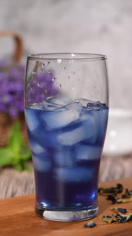 Milk slowly pours from a small white pitcher into iced butterfly pea tea, creating soft white swirls and turning the drink a pale blue. Part 4 of the series, Vertical video. Real time.