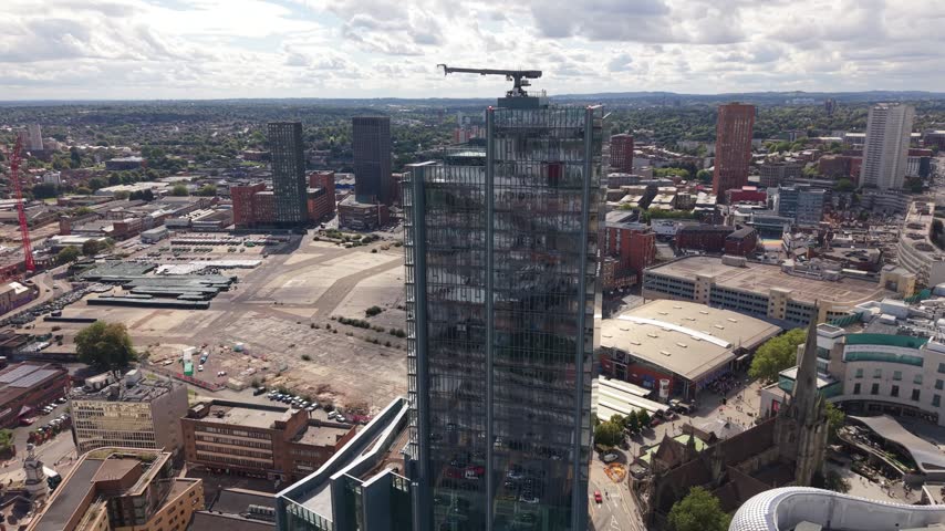 Aerial view of a tall modern glass skyscraper with a dark reflective facade rising above Birmingham city centre, surrounded by contemporary buildings and dense urban development.
