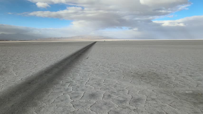 Dark tire tracks cut across the cracked surface of Salinas Grandes salt flat under wide clouds and blue sky, useful as an abstract travel or desert landscape background.