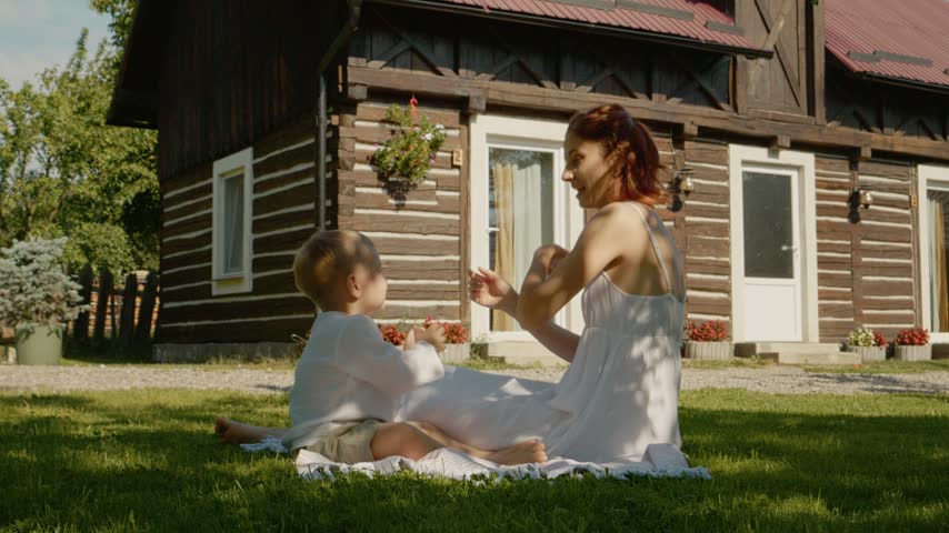 A mother sits on the grass with her child in front of a traditional house in Bucovina. They play together while enjoying the warm sunlight and fresh air.
