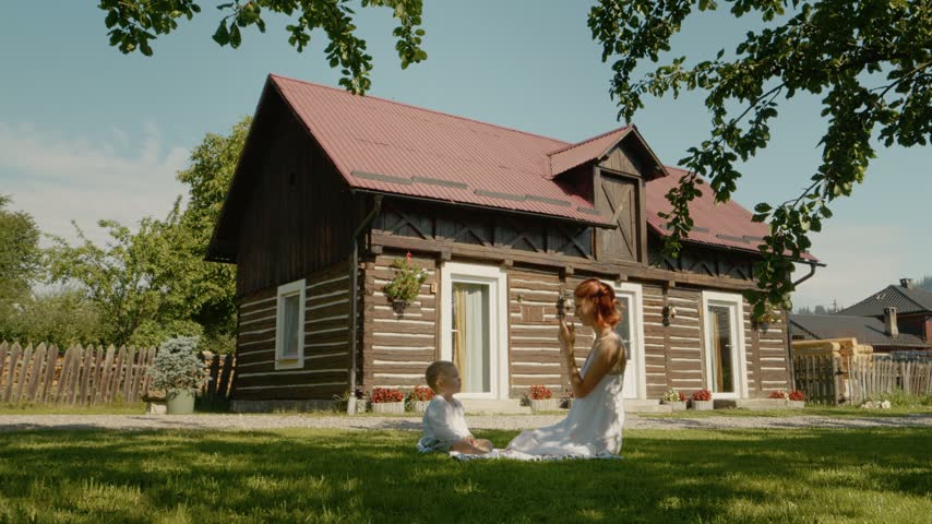 A mother shares fun moments with her child on the grass near a traditional house in Bucovina. They enjoy games and activities while sitting outside.
