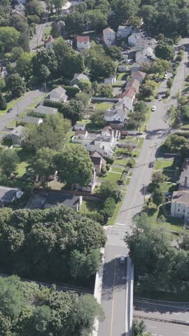 A vertical aerial pan moves from a road bridge toward detached homes and a curving junction in White Plains, New York, revealing summer tree cover, green yards and the pattern of suburban streets.
