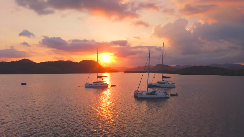 Aerial sunset shot of sailboats anchored in Coron Bay with glowing orange reflections and tropical island silhouettes.