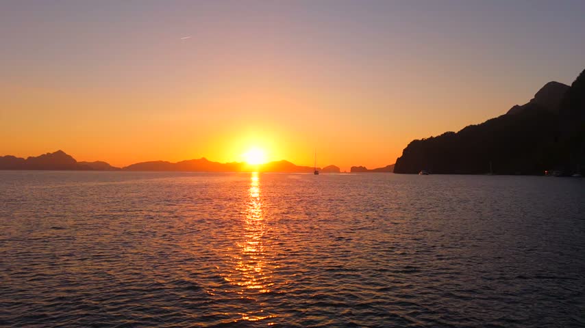 Bright orange sunset over calm water in Coron Bay with distant sailboat and dramatic limestone cliffs in silhouette.