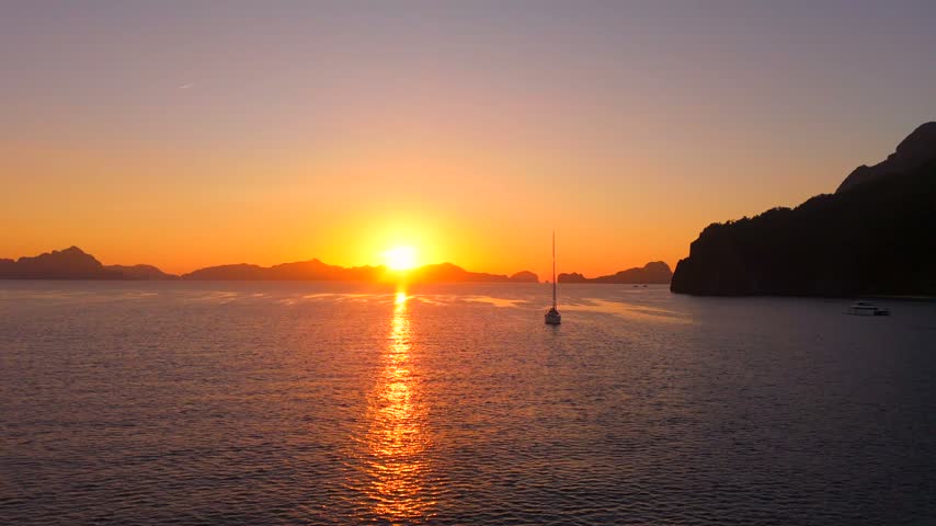 Wide sunset seascape in Coron Bay with a small sailboat, orange sky and glowing reflection across calm water.