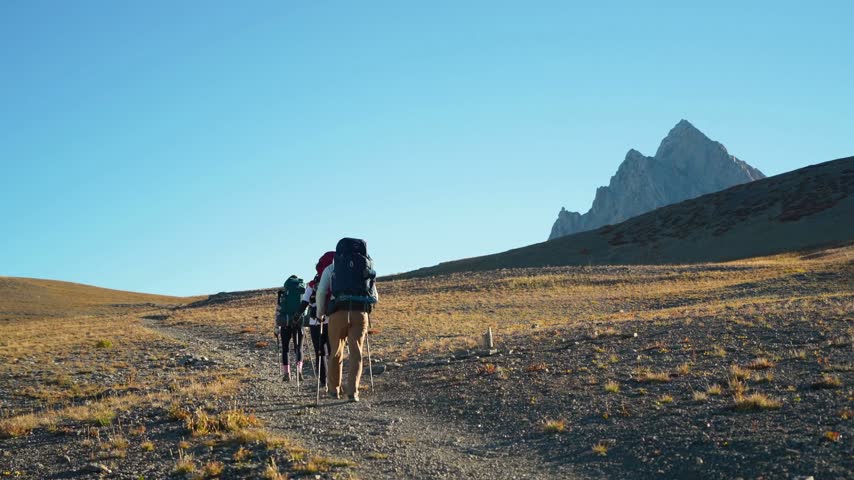 Group of hikers journeys along a dirt trail surrounded by grassy hills and rocky mountains. Bright blue sky adds depth to the serene outdoor environment.