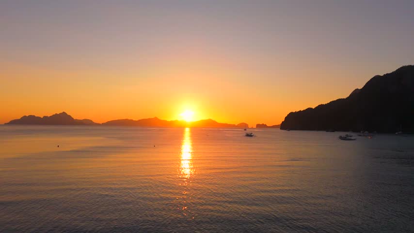 Wide view of a glowing orange sunset over Coron Bay with distant boats, calm sea and limestone island silhouettes.
