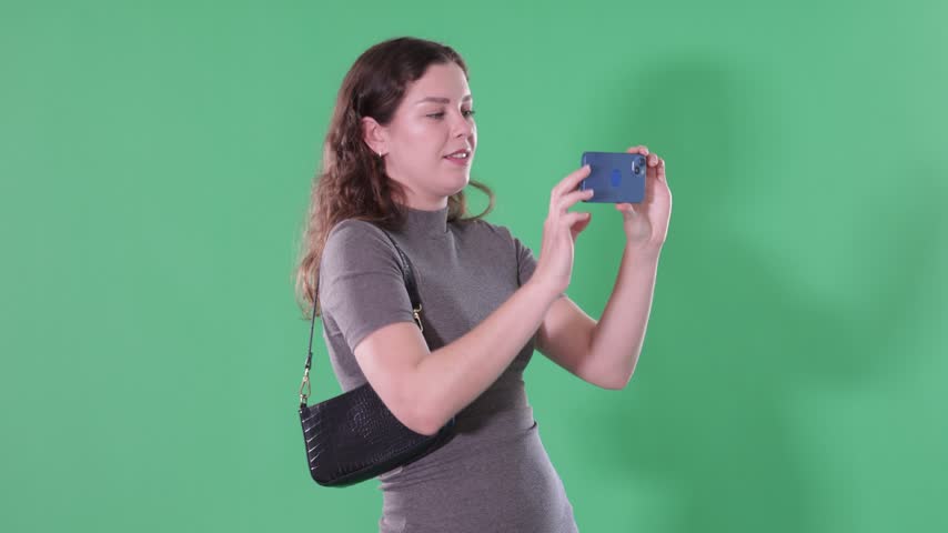 Young woman stands against green screen, holding smartphone up to take photo or scan QR code 