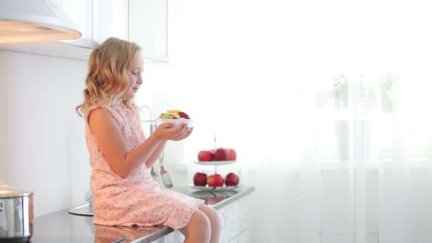 Little girl sitting on table and holding plate of fruit yoghurt
