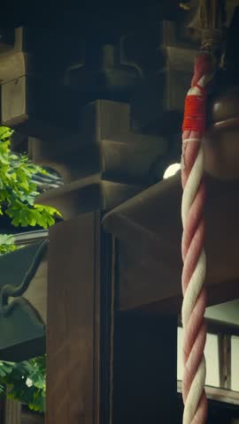 cinematic vertical tilt-down shot of a traditional Japanese bronze shrine bell and thick red and white rope hanging from a wooden roof