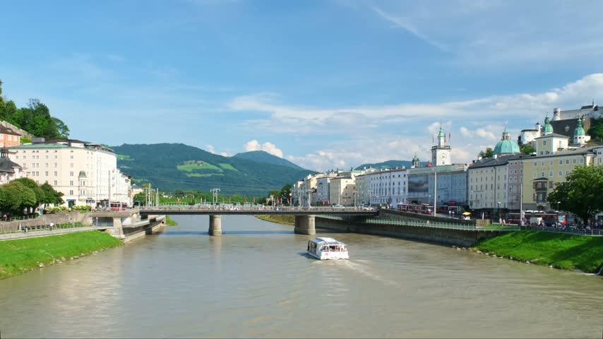 A sightseeing boat passes on the Salzach River through historic Salzburg, Austria, with a fortress on the hill