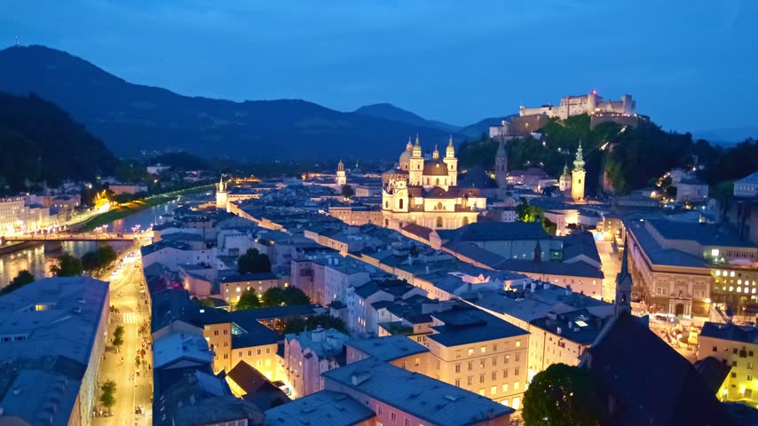 Wide aerial view of Salzburg at twilight with the Salzach River, historic Old Town buildings, and Hohensalzburg Fortress
