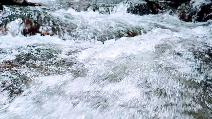 Close-up HD slow-motion footage of rushing water splashing and flowing over natural rocks in a large rocky stream. Detailed water movement, foam, and droplets create