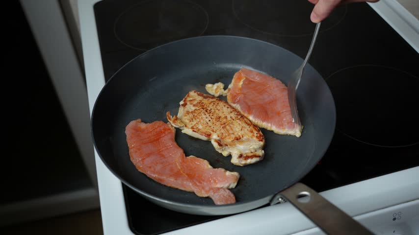 Turning Two Turkey Steaks in a Frying Pan. Two turkey steaks are turned over in a hot frying pan while cooking on the stovetop.