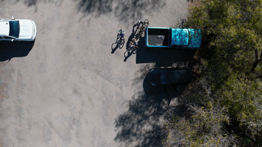 Three mountain bikers ready their bikes beside a pickup at a gravel trailhead at Gamehaven, Rochester, Minnesota, tree shadows falling across the parking area. Aerial top down