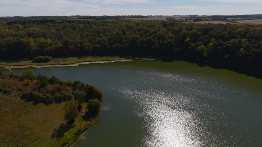 Sunlight glitters on Gamehaven Reservoir at Rochester, Minnesota as the camera climbs, the earth dam and mown meadows opening out below wooded bluffs and farm fields. Aerial rising shot