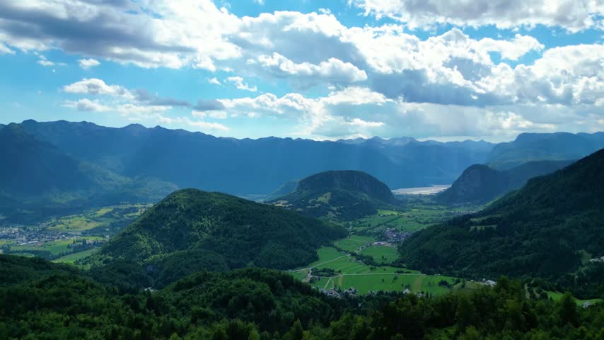 Panoramic view of lush green valleys and majestic Julian Alps surrounding Lake Bohinj, Slovenia. Dynamic clouds cast shadows over the serene landscape.