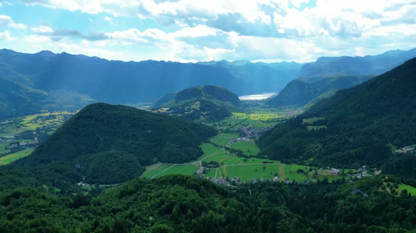 Panoramic view of lush green valleys and majestic Julian Alps surrounding Lake Bohinj, Slovenia. Dynamic clouds cast shadows over the serene landscape.