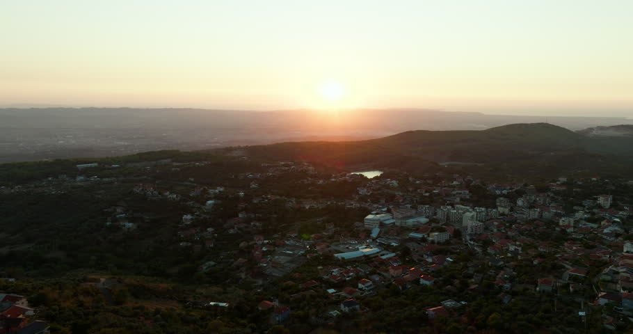 The sun sets beyond Kruje in Albania as an aerial view passes over the town and castle grounds. Red rooftops, wooded hills and distant plains fill the scene.