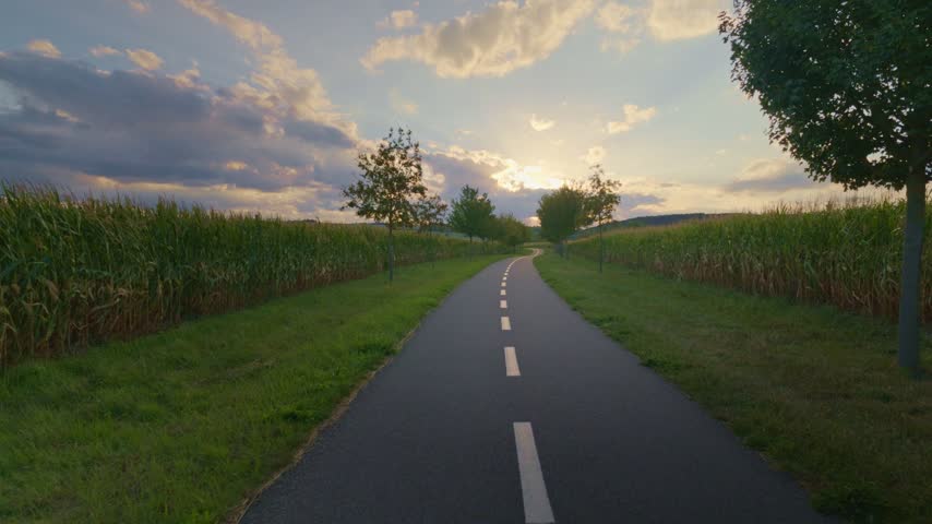 Forward motion along a straight road bordered by trees and cornfields.
Sunset light creates a peaceful rural landscape.