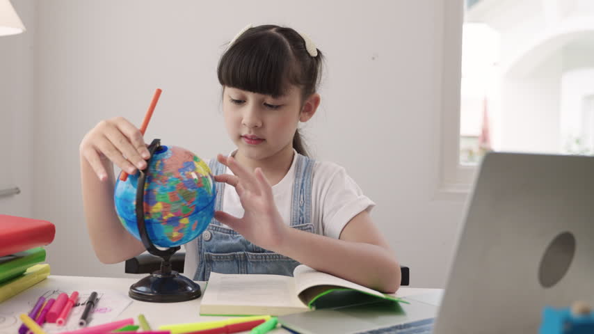Cute Asian schoolgirl sitting at a desk using laptop computer for studying online learning via video calling and a remote teacher while having a geography lesson with a globe model in homeschool room. 