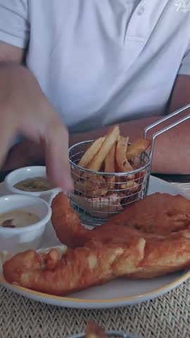 A hand squeezing lime all over a battered fish fillet served with fried potatoes, a classic british fish and chips