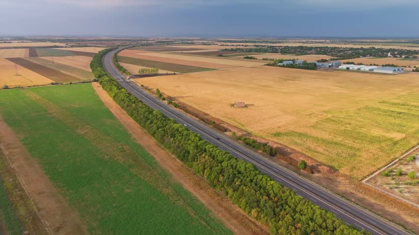 Iváncsa, Hungary, M6 motorway with traffic and overpass through rural farmland, grain fields and industrial buildings under a cloudy sky, aerial view.