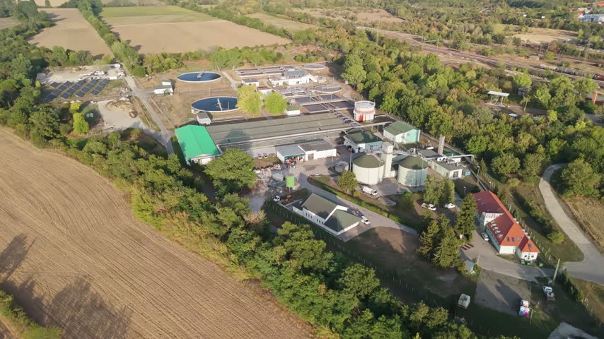 Veszprém, Hungary sewage treatment plant with circular clarifiers and water tanks amid rural fields and forests, municipal wastewater facility and biogas plant in aerial view.