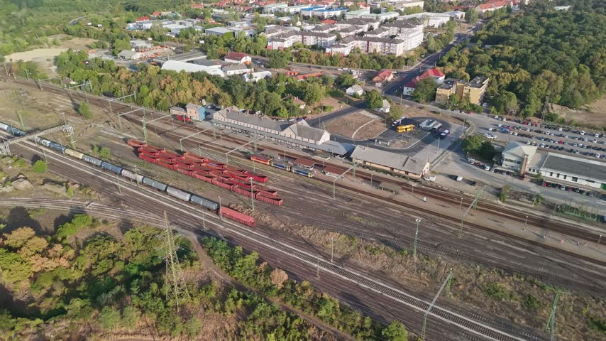 Veszprém, Hungary railway station with passenger and freight trains, rail yard, bus station and urban infrastructure among greenery, captured in an aerial view.
