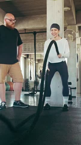 Smiling woman performs a battle rope workout while a male fitness coach watches at the gym, showing strength, endurance, motivation and energetic functional training.