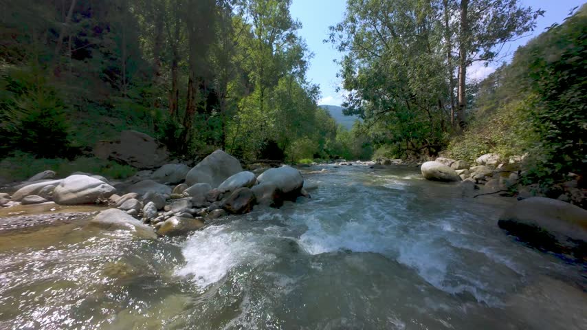 Low-altitude drone flight over a rocky alpine river flowing through a lush green forest under a clear sky.