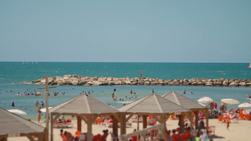 Vibrant summer day at a crowded beach in Tel Aviv, Israel, with people swimming in the Mediterranean Sea, relaxing under gazebos, and enjoying the sunny weather by the breakwater