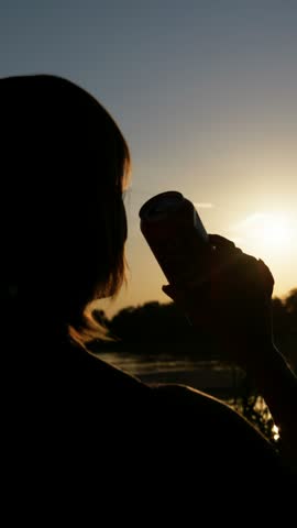 Silhouette of a person raising a beverage can against glowing sunset sky near a lake. Backlit composition highlights outdoor summer refreshment for mobile lifestyle campaigns.