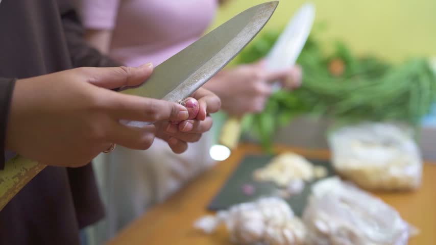 Close view of hands trimming small purple onion with kitchen knife over warm wooden table, soft indoor light, blurred helper, and leafy produce in background.