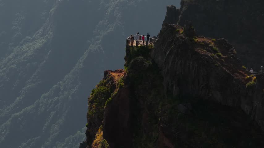 Tourists stand on a dramatic mountain viewpoint above clouds along the PR1 trail, rugged peaks and lush slopes highlighted by afternoon sunlight in this dynamic wide-angle landscape.