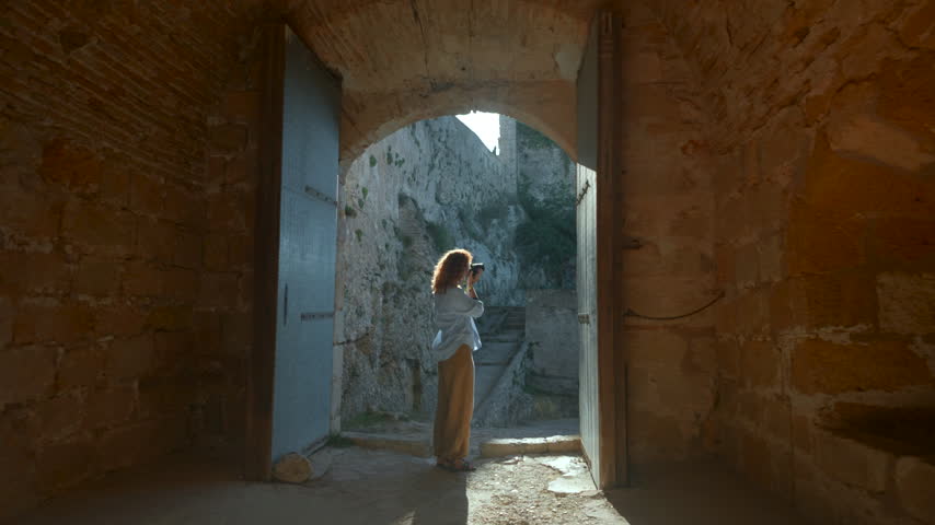 Young woman photographing sunlit fortress entrance from shadowed stone gateway with tall arch, weathered brick walls, heavy doors, centered framing, and dramatic backlight