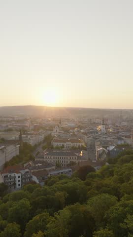 Sun sets over central Brno as church spires, historic rooftops and a dense green park fill the hazy city skyline.