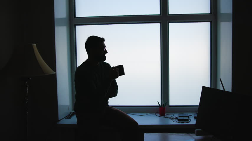Silhouette of seated bearded man holding mug on windowsill beside bright four-pane frosted window with soft backlit shadows