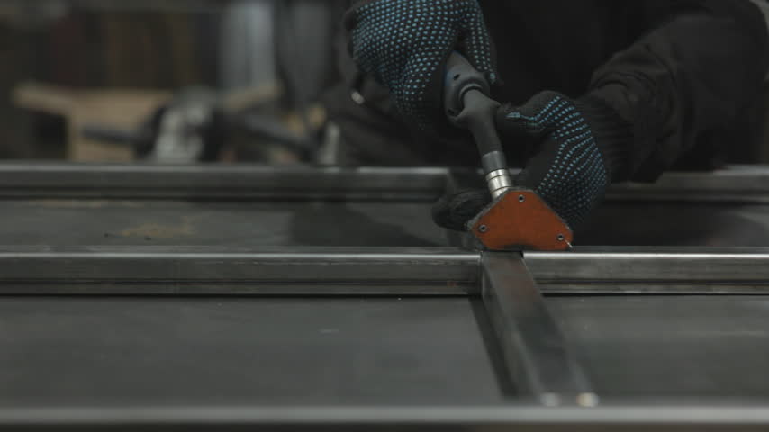 Gloved worker guides handheld cutting tool across steel in workshop, producing bright sparks and cool blue light over reflective metal bars during metal fabrication