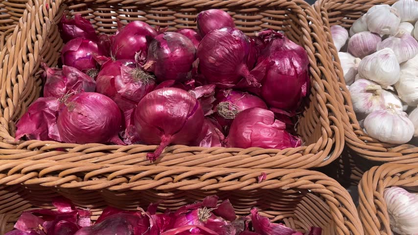 A horizontal view across woven produce baskets containing purple onions, white winter garlic and long carrots with visible soil residue. Natural vegetable market display highlighting fresh agricultural produce and rustic food textures.