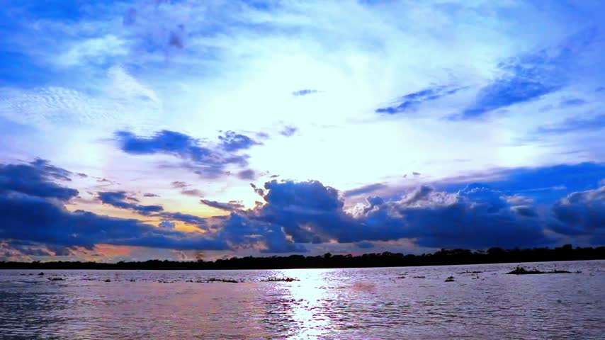 Scenic wide shot of a tranquil river with water reflection under a vibrant blue sky filled with dramatic clouds and soft sunlight during sunset.