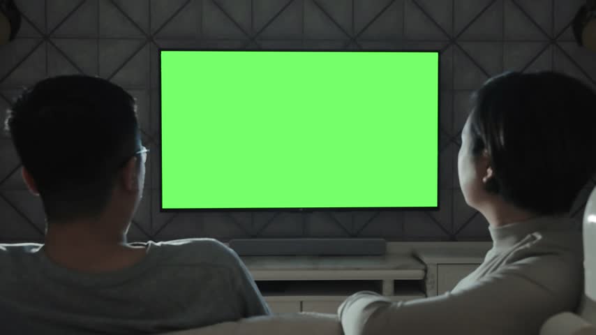 This video captures a pair of individuals seated on a couch in a dim living room, focused intently on a large television screen featuring a vibrant green chroma key background