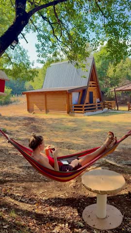 Woman drinks coffee while reclining in a hammock under trees and looking toward a wooden cabin on a sunny day.