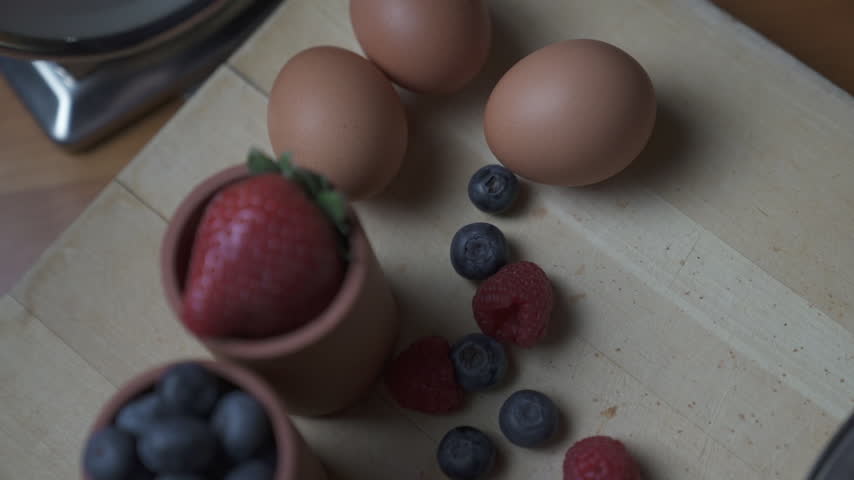 The camera tilts down over a fresh strawberry, blueberries and raspberries in terracotta cups on a wooden cutting board, with eggs and loose berries scattered around, ready for baking