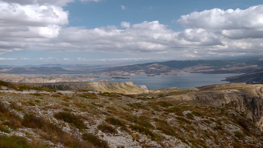 Panoramic rocky hillside overlooking broad reservoir, layered mountains, drifting clouds, dry grasses, rugged cliffs, and remote open terrain under soft daylight