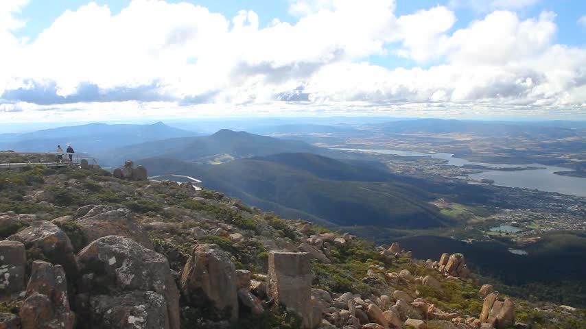 Panoramic aerial view from the summit of Mount Wellington kunanyi overlooking Hobart, Tasmania, with the Derwent River, city sprawl, and forested hills under a partly cloudy sky.